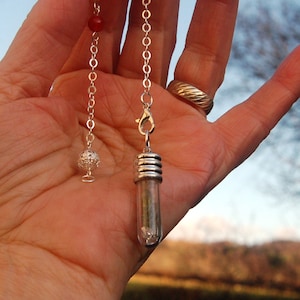 Puede incluir: Una cadena de plata con un péndulo de cristal transparente y una bola de plata en el extremo. La cadena se sostiene en una mano con un anillo en el dedo.