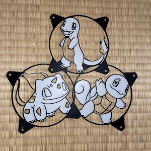 May include: Three black metal fan-shaped designs, each with a white and light gray cartoon character inside. The characters are a lizard, a turtle, and a plant-like creature. The background is a light brown woven mat.