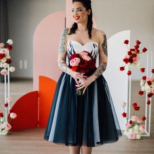 Könnte beinhalten: Ein trägerloses, knielanges Kleid mit einem schwarz-weißen Oberteil und einem schwarzen Tüllrock. Die Person im Bild hält einen Strauß roter und rosa Rosen und trägt rote Absätze. Der Hintergrund zeigt Blumenarrangements.