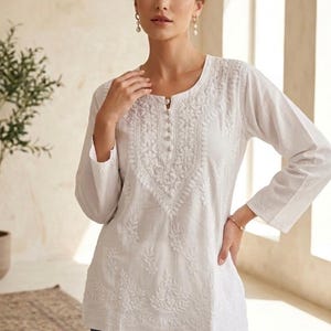 Op de afbeelding: Witte geborduurde tuniek met lange mouwen en een ronde halslijn. De tuniek heeft gedetailleerde bloemborduurwerk rond de halslijn en op de voorkant. De merknaam "Jeehva" is linksboven zichtbaar. Het model draagt een blauwe jeans.