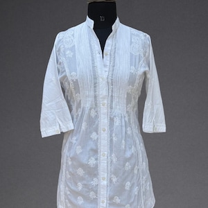 May include: White tunic dress with detailed embroidery and a mandarin collar. The dress has a button-down front, three-quarter sleeves, and a gently flared hem. The Jeehva logo is visible in the upper right corner.