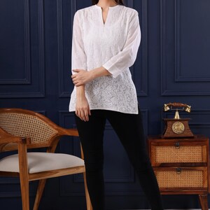 May include: A woman wearing a white embroidered tunic and black pants. The tunic has a V-neck and three-quarter sleeves. The image includes the text "Jeehva Threads that breathe tradition".