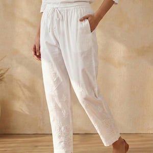 Può includere: Pantaloni bianchi ricamati con vita con coulisse e tasche. I pantaloni presentano ricami floreali lungo l'orlo. La modella indossa ballerine in pelle marrone. L'outfit è realizzato in tessuto leggero.
