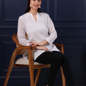 May include: A woman wearing a white embroidered tunic with a V-neck, paired with black pants, seated on a wooden chair. The tunic features detailed floral embroidery. The image includes the text "Jeehva Threads that breathe tradition".