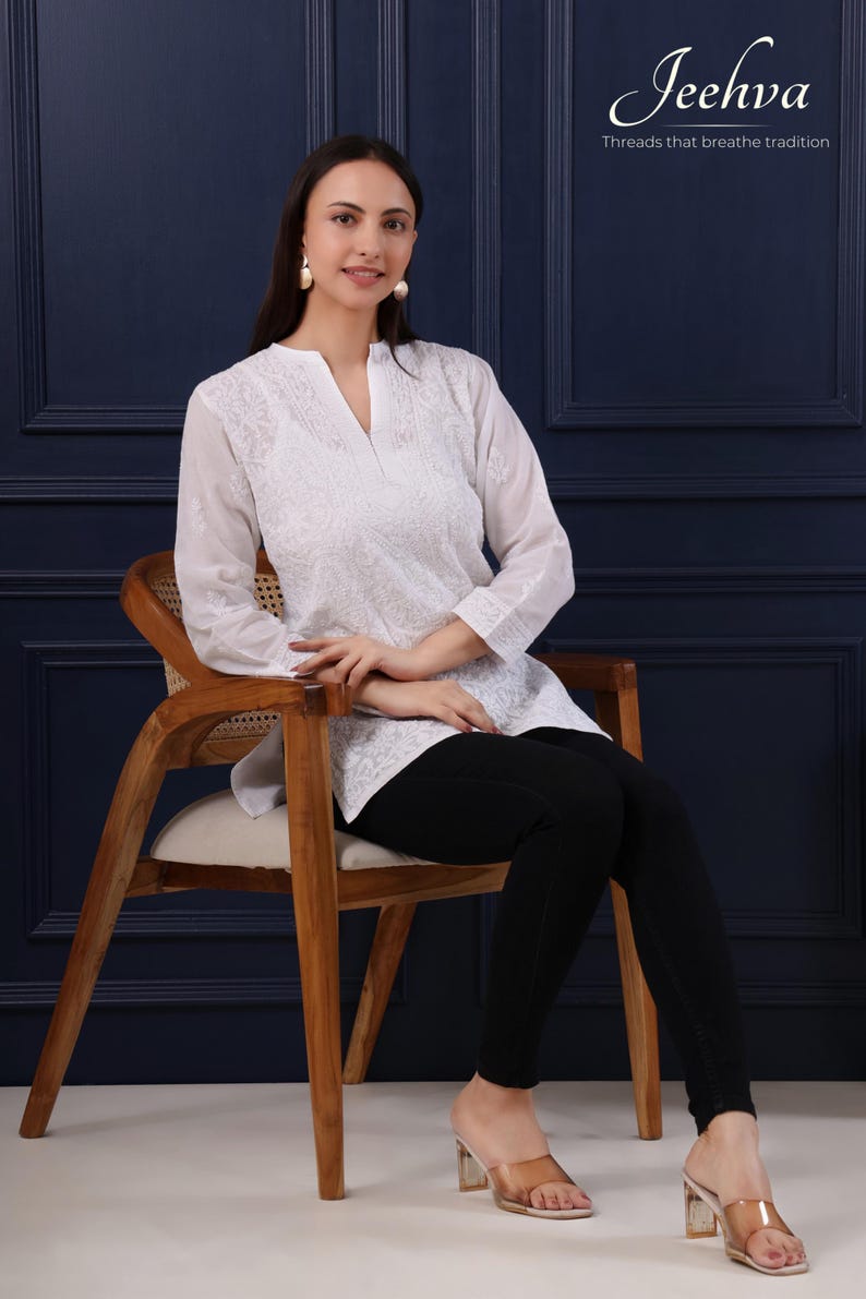 May include: A woman wearing a white embroidered tunic with a V-neck, paired with black pants, seated on a wooden chair. The tunic features detailed floral embroidery. The image includes the text "Jeehva Threads that breathe tradition".