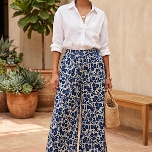 May include: A white long-sleeved button-down shirt with a collar and a chest pocket paired with wide-leg trousers featuring a blue and white floral pattern. The outfit includes a small woven straw handbag and tan wedge sandals.