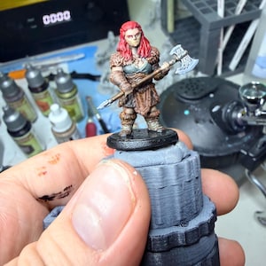 May include: A miniature figurine of a warrior with bright red hair, wearing brown fur-trimmed armour and a grey chest piece. The figure holds a double-headed battle axe with a silver blade and a long wooden handle, standing on a black circular base.