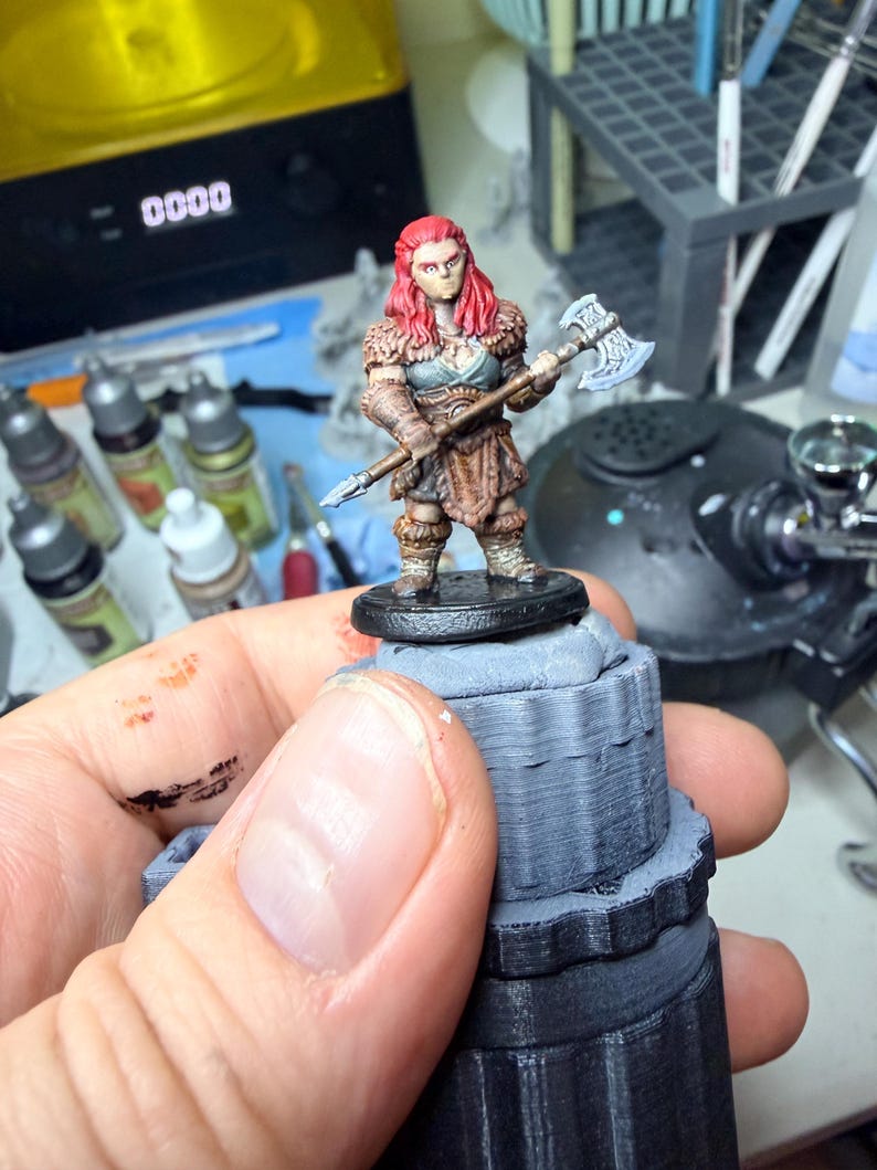 May include: A miniature figurine of a warrior with bright red hair, wearing brown fur-trimmed armour and a grey chest piece. The figure holds a double-headed battle axe with a silver blade and a long wooden handle, standing on a black circular base.