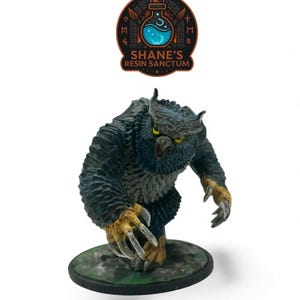 May include: A painted miniature figurine of an owlbear creature with dark blue-black feathers, yellow eyes, and tan-coloured claws, standing on a circular base. Above the figure is a logo reading SHANE'S RESIN SANCTUM with a potion bottle.