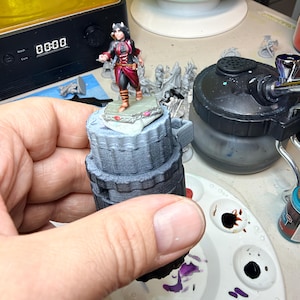 May include: A miniature figurine of a character with dark hair and horns, wearing a black and red outfit with brown boots, standing on a hexagonal base. The figure is mounted on a grey, cylindrical, 3D-printed handle for painting.