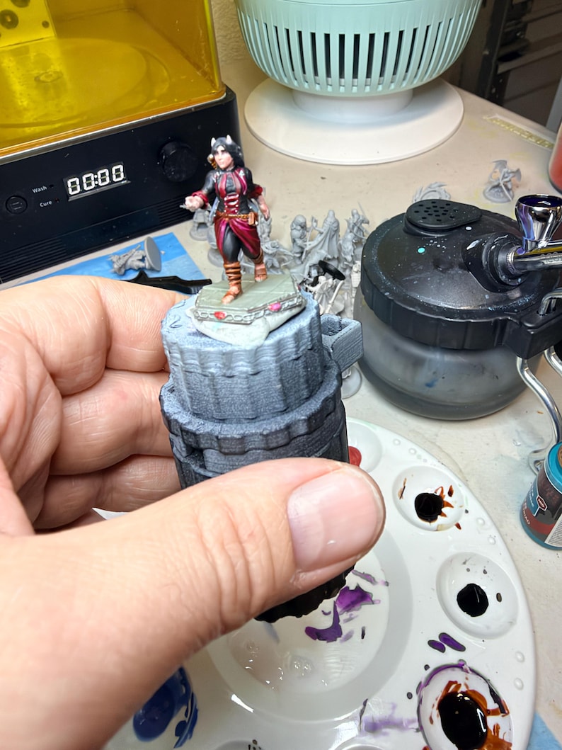 May include: A miniature figurine of a character with dark hair and horns, wearing a black and red outfit with brown boots, standing on a hexagonal base. The figure is mounted on a grey, cylindrical, 3D-printed handle for painting.