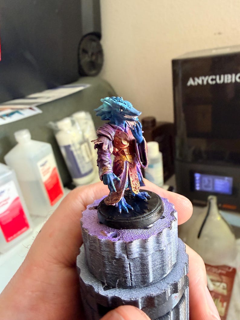 May include: A small, hand-painted fantasy figurine featuring a blue-skinned reptilian creature with a long snout. The figure wears a layered purple and gold robe with intricate detailing and stands on a circular black base.