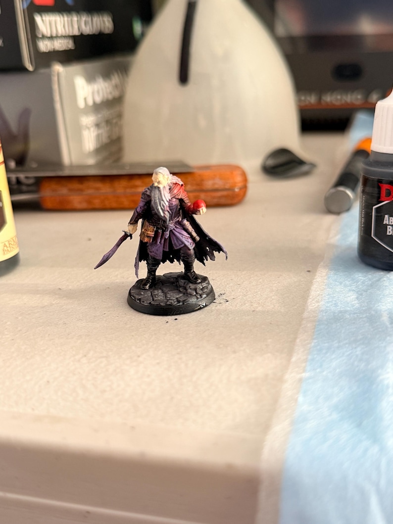 May include: A miniature figurine of a bearded character with long white hair, wearing a purple tunic, dark cloak, and black boots. The figure holds a small sword in one hand and a red sphere in the other, standing on a circular stone base.