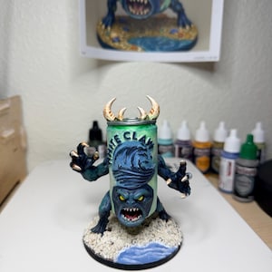 May include: A hand-painted miniature figurine of a monster with a White Claw hard seltzer can for a torso. The creature has dark blue skin, sharp claws, yellow eyes, and a wide mouth with teeth. It stands on a circular base with sand and water.