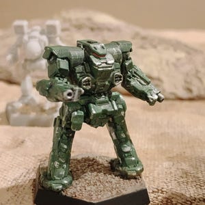 May include: A painted green miniature robot figurine with metallic silver highlights on the torso and limbs. The bipedal mech features a blocky design, a central cockpit area, and weapon ports on the chest, standing on a hexagonal base.