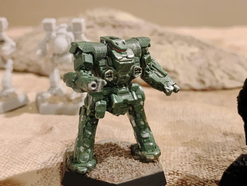 May include: A painted green miniature robot figurine with metallic silver highlights on the torso and limbs. The bipedal mech features a blocky design, a central cockpit area, and weapon ports on the chest, standing on a hexagonal base.