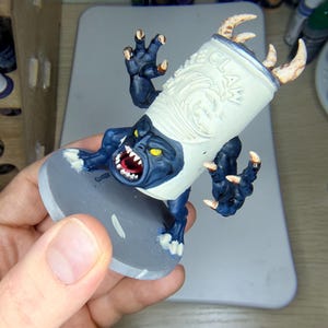 May include: A painted miniature figurine featuring a dark blue creature with yellow eyes and sharp teeth emerging from a white cylindrical can labelled CLAW. The figure has clawed limbs, tan horns, and is mounted on a circular grey base.