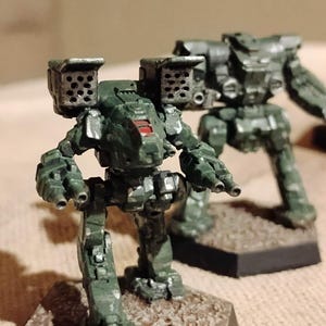 May include: Two miniature dark green robot figurines standing on hexagonal bases. The front robot features dual shoulder-mounted missile launchers with visible circular ports and a red cockpit window, while the second robot stands behind.