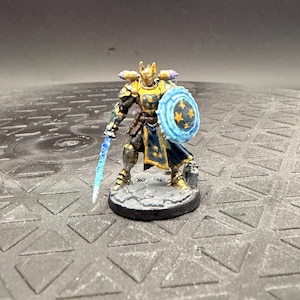 May include: A miniature figurine of a warrior in gold and dark blue armour, holding a translucent blue sword and a round shield with a blue energy effect and yellow star patterns, standing on a circular grey stone-textured base.