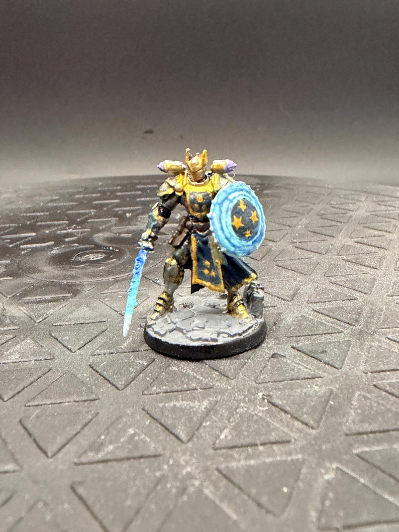 May include: A miniature figurine of a warrior in gold and dark blue armour, holding a translucent blue sword and a round shield with a blue energy effect and yellow star patterns, standing on a circular grey stone-textured base.