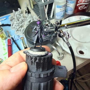 May include: A miniature figurine of a humanoid character with purple skin and small horns, wearing a dark coat and scarf, standing on a circular base. The figure is held in place by a black 3D-printed handle while being painted.