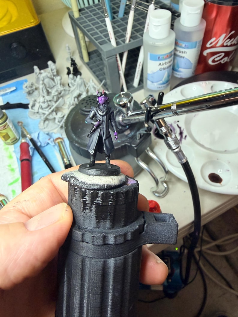 May include: A miniature figurine of a humanoid character with purple skin and small horns, wearing a dark coat and scarf, standing on a circular base. The figure is held in place by a black 3D-printed handle while being painted.