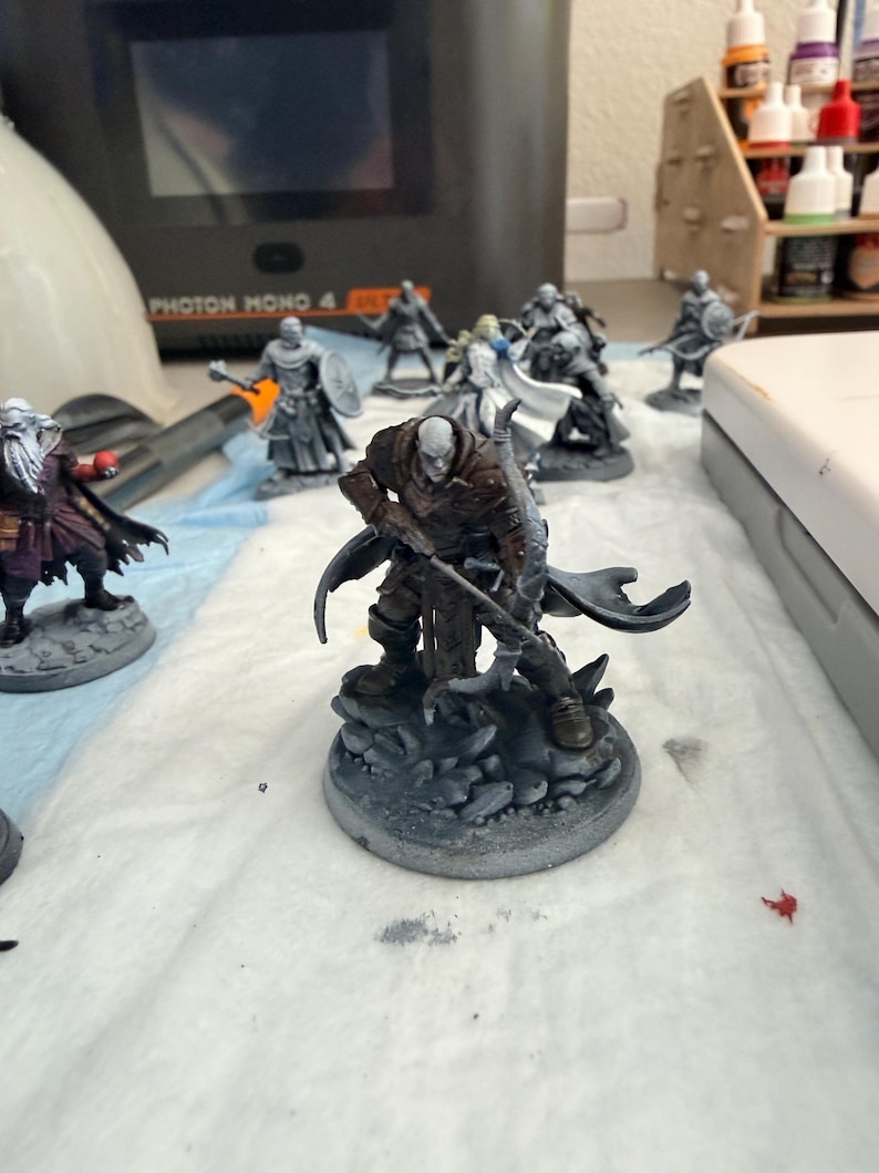 May include: A miniature fantasy figure of a bald, pale-skinned warrior wearing brown armour and a dark cloak, holding a bow and standing on a rocky base. The figure is part of a collection of unpainted and partially painted gaming miniatures.