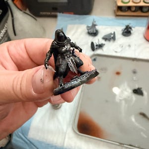 May include: A miniature tabletop gaming figure of a hooded character wearing dark armour with silver highlights, standing on a circular base. The figure features a cloak, detailed chest plating, and a weapon held in one hand, painted in shades of black, grey, and metallic silver.