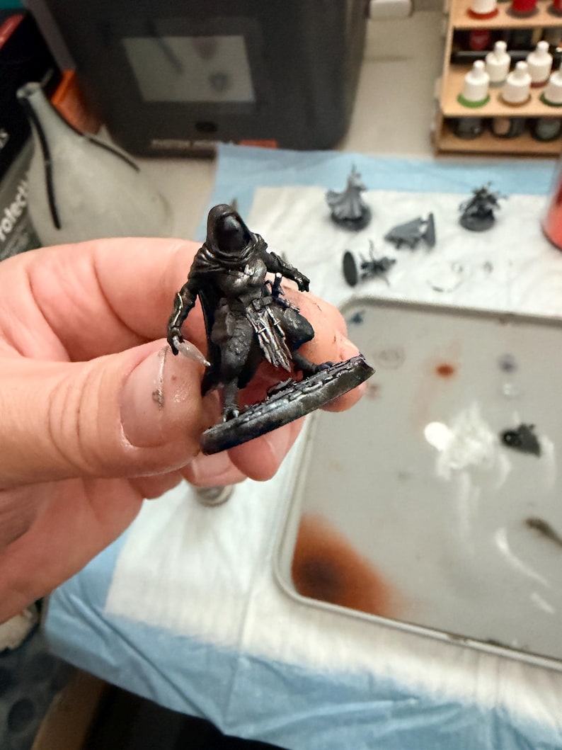 May include: A miniature tabletop gaming figure of a hooded character wearing dark armour with silver highlights, standing on a circular base. The figure features a cloak, detailed chest plating, and a weapon held in one hand, painted in shades of black, grey, and metallic silver.