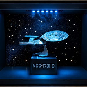 Könnte beinhalten: Ein Modell des Raumschiffs Enterprise NCC-1701-D in einem schwarzen Schattenkastenrahmen. Das Schiff ist auf einem Ständer auf einem schwarzen Sockel mit der Aufschrift NCC-1701-D montiert. Der Hintergrund zeigt ein Sternenfeld mit blauer LED-Beleuchtung.