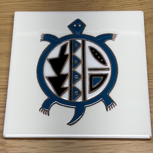 May include: Square white ceramic tile featuring a blue turtle design with black and brown accents. The turtle's shell is filled with geometric shapes. The tile is likely used as a coaster or decorative piece.