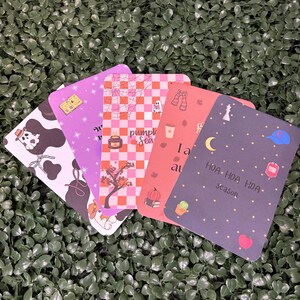 Puede incluir: Cinco tarjetas rectangulares con esquinas redondeadas que presentan varios diseños: un estampado de vaca en blanco y negro con un fantasma, una tarjeta morada con estrellas, una tarjeta a cuadros rosa y naranja con el texto pumpkin season, una tarjeta coral y una gris oscuro.