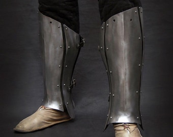 May include: Steel leg armour, riveted and curved to fit the legs, with leather straps and buckles. The armour is worn over black padded trousers and light brown leather shoes. The image is set against a dark background.