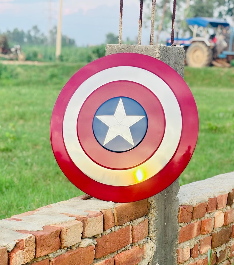 K&ouml;nnte beinhalten: Runder Schild in Rot, Wei&szlig; und Blau mit einem wei&szlig;en Stern in der Mitte, &auml;hnlich dem Schild von Captain America. Der Schild hat eine metallische Oberfl&auml;che und ist auf einer Ziegelmauer platziert.