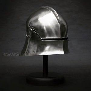 Può includere: Un elmo medievale lucido, color argento, con la parte superiore arrotondata e una visiera. L'elmo è esposto su un supporto nero su sfondo scuro. Il testo "IronArtisanGifts" è visibile.
