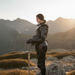 May include: A person in black armor stands on a rocky hillside, holding a long sword. The armor covers the shoulders, arms, and legs. The sword has a detailed hilt and a long, silver blade. The background features mountains and a sunset.