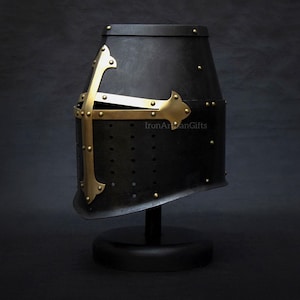May include: A black medieval helmet with a brass cross design. The helmet is displayed on a black stand against a dark background. The helmet has small rivets and a slightly angled shape.