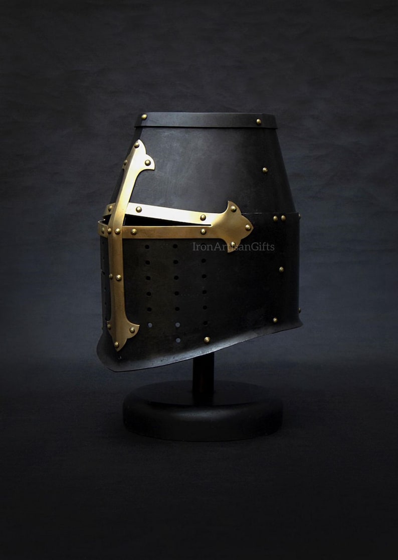 May include: A black medieval helmet with a brass cross design. The helmet is displayed on a black stand against a dark background. The helmet has small rivets and a slightly angled shape.