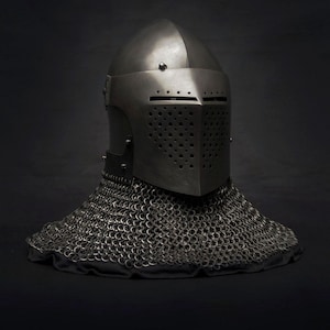 Puede incluir: Un yelmo medieval de caballero con construcción de acero remachado. El yelmo presenta una visera con ranuras horizontales y una placa frontal perforada. Una cofia de cota de malla se extiende desde la base, ofreciendo protección para el cuello.