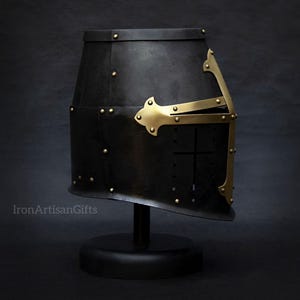 May include: Black medieval helmet with brass accents, rivets, and a cross design. The helmet is displayed on a black stand against a dark background. The helmet has a distinctive shape and is likely made of metal.