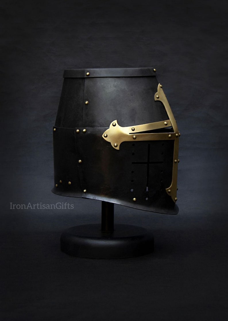 May include: Black medieval helmet with brass accents, rivets, and a cross design. The helmet is displayed on a black stand against a dark background. The helmet has a distinctive shape and is likely made of metal.