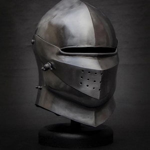 May include: A polished, silver-colored medieval helmet with a visor and riveted details. The helmet is displayed on a black stand against a dark background, showcasing its historical design and craftsmanship.