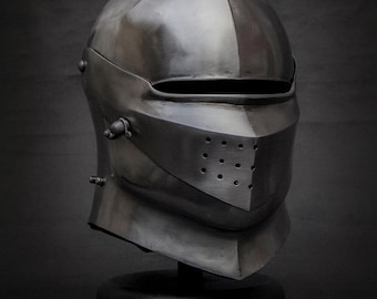 May include: A polished, silver-colored medieval helmet with a visor and riveted details. The helmet is displayed on a black stand against a dark background, showcasing its historical design and craftsmanship.
