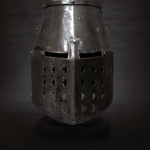 Peut inclure: Casque en métal vieilli, avec un dessus plat et une face carrée. L'avant présente des rangées de trous d'aération en forme de croix. Le casque est posé sur une base sombre et ronde, sur fond sombre.