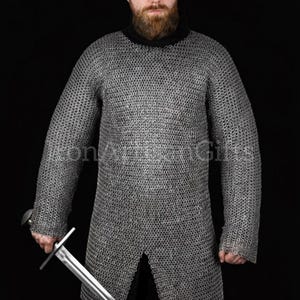 Puede incluir: Una camisa de cota de malla plateada con mangas largas y una abertura frontal. La camisa se usa sobre una prenda negra. Una persona sostiene una espada plateada con una empuñadura en forma de cruz. El fondo es negro.