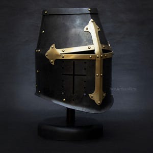 May include: A black metal knight's helmet with a gold-colored cross design. The helmet is mounted on a black stand against a dark background. The cross is riveted to the helmet. The text "IronArtisanGifts" is visible.