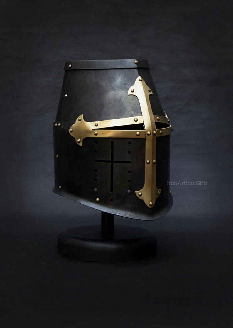 May include: A black metal knight's helmet with a gold-colored cross design. The helmet is mounted on a black stand against a dark background. The cross is riveted to the helmet. The text "IronArtisanGifts" is visible.