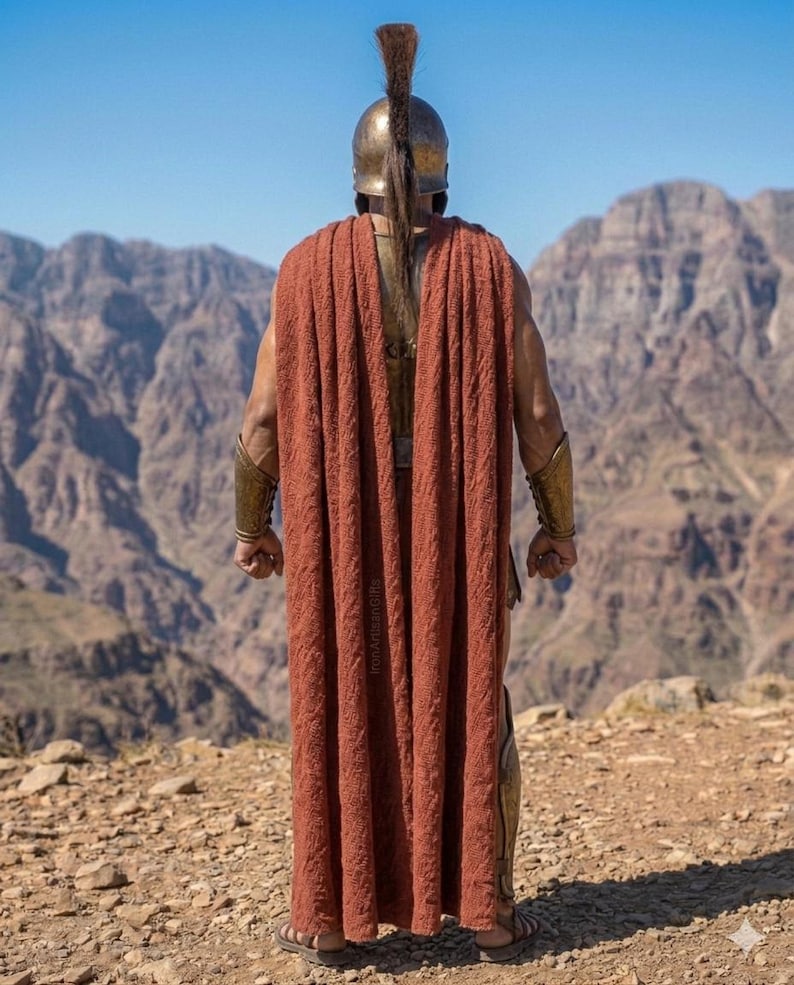 May include: A person wearing a bronze helmet with a tall plume and a rust-colored cape, standing with their back to the viewer. The person is wearing bronze arm and leg armor. Mountains and a blue sky are in the background.