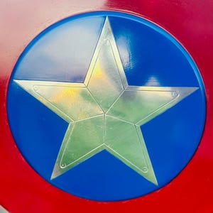 K&ouml;nnte beinhalten: Ein rot-wei&szlig;-blauer Schild mit einem silbernen Stern in der Mitte. Der Schild hat einen roten Au&szlig;enring, einen blauen Innenkreis und einen silbernen, f&uuml;nfzackigen Stern in der Mitte. Der Stern ist metallisch und hat eine strukturierte Oberfl&auml;che.