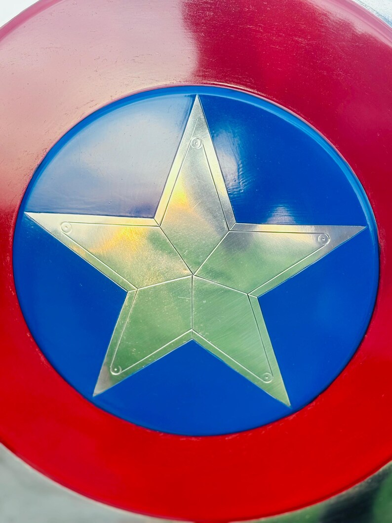 K&ouml;nnte beinhalten: Ein rot-wei&szlig;-blauer Schild mit einem silbernen Stern in der Mitte. Der Schild hat einen roten Au&szlig;enring, einen blauen Innenkreis und einen silbernen, f&uuml;nfzackigen Stern in der Mitte. Der Stern ist metallisch und hat eine strukturierte Oberfl&auml;che.